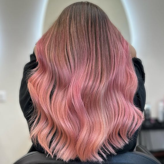 Back of model’s head with long, wavy, dirty blonde hair featuring a rose gold and pink balayage.