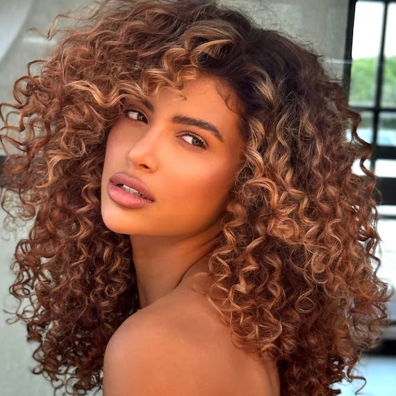 Model with long, dark brown, curly hair featuring golden, sun-kissed highlights.