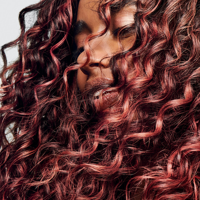 Close-up of model with dark brown, curly hair featuring coral highlights.