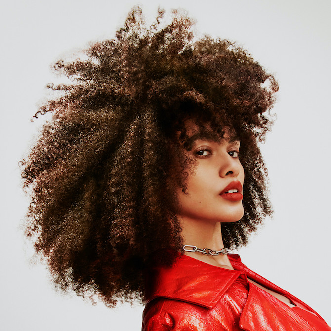 Headshot of a model with very curly brown hair wearing a red jacket and red lipstick