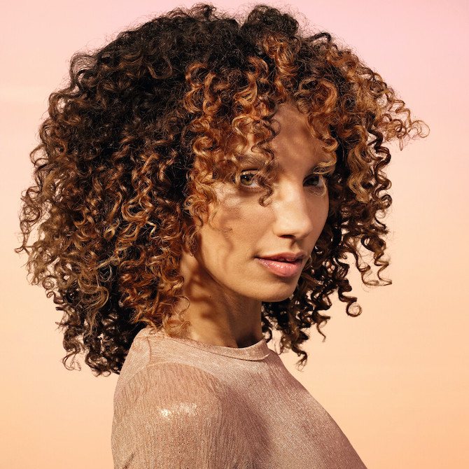 Model with long, voluminous, curly, brown hair featuring bronze highlights.
