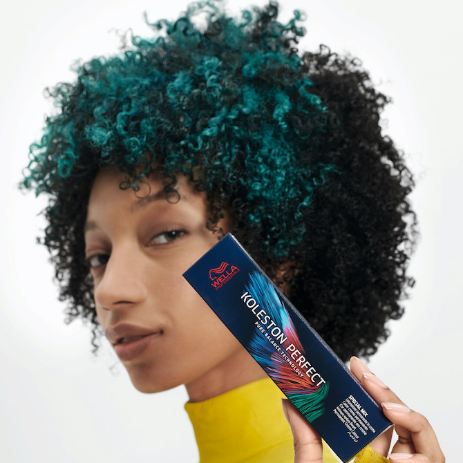 Model with curly, black hair and teal highlights holds up a box of Koleston Perfect permanent hair colour.