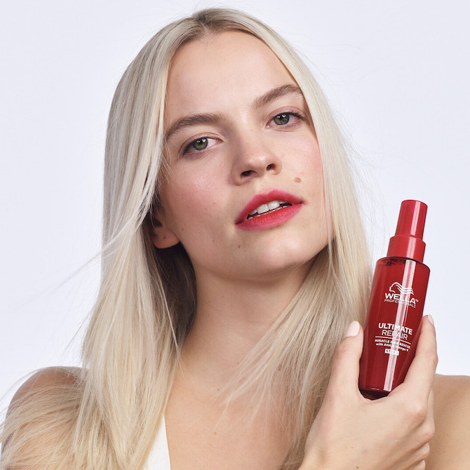 Model with platinum blonde hair and red lips holds a red bottle of Ultimate Repair Miracle Hair Rescue by Wella Professionals 