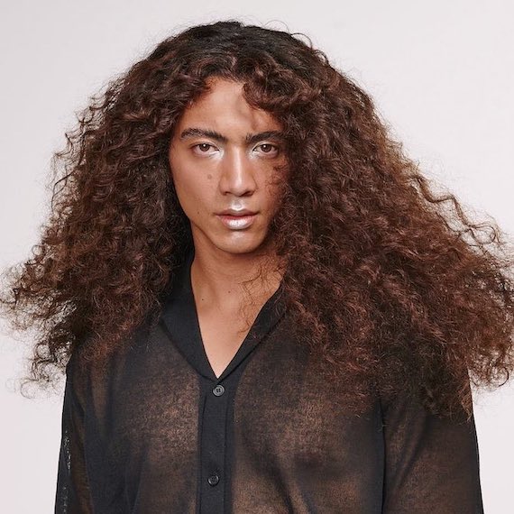 Headshot of a person with long curly chocolate brown hair wearing a black sheer shirt