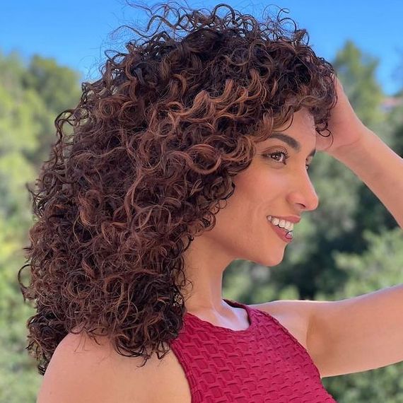 Side profile of woman with dark brown curly hair. She stands outside in blue sky surrounded by trees. 