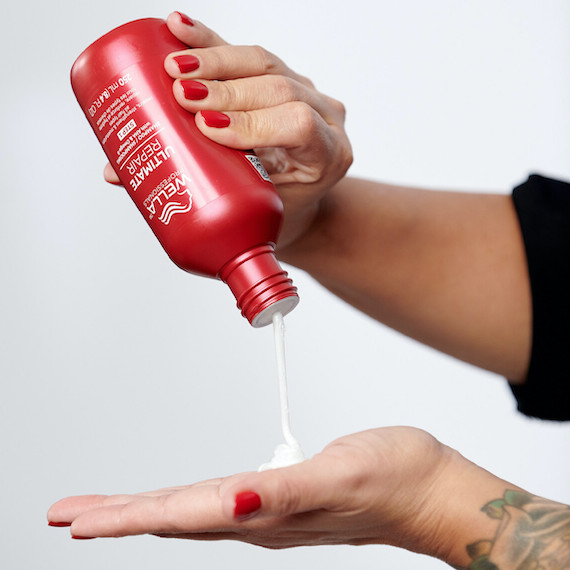 Nahaufnahme von Händen mit rot lackierten Nägeln, die eine Flasche Ultimate Repair Shampoo drücken