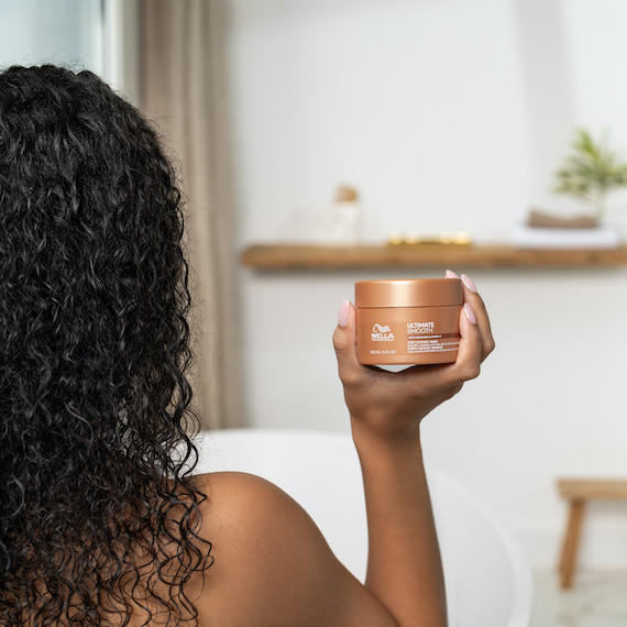 Model with long, dark, curly hair holds up a tub of ULTIMATE SMOOTH Mask.