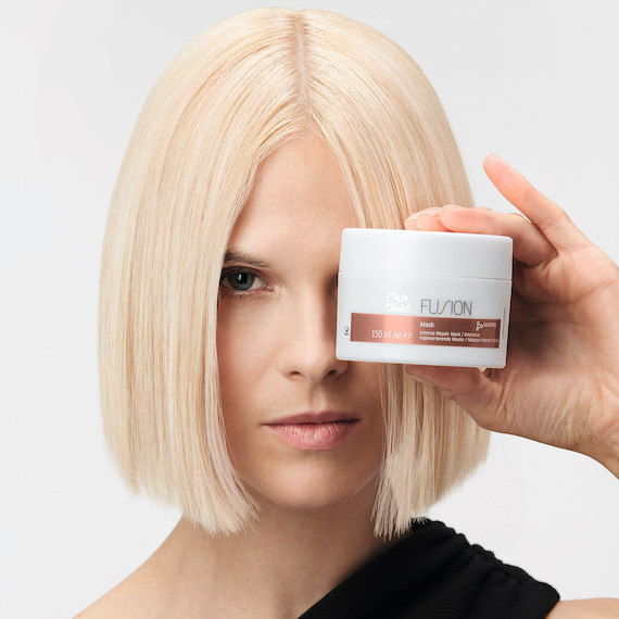 Model with short, platinum blonde hair holds the Fusion Intense Repair Hair Mask over their eye