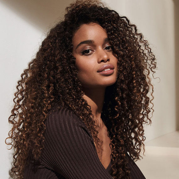 Model with long, voluminous curly hair, featuring dark brown roots and caramel brown ends.
