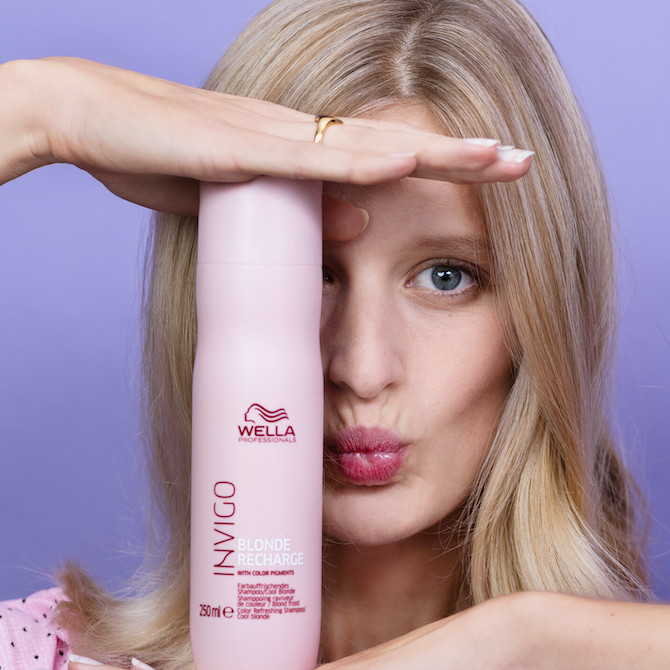 Model with long, blonde hair holds a bottle of Wella INVIGO Blonde Recharge in front of their face.