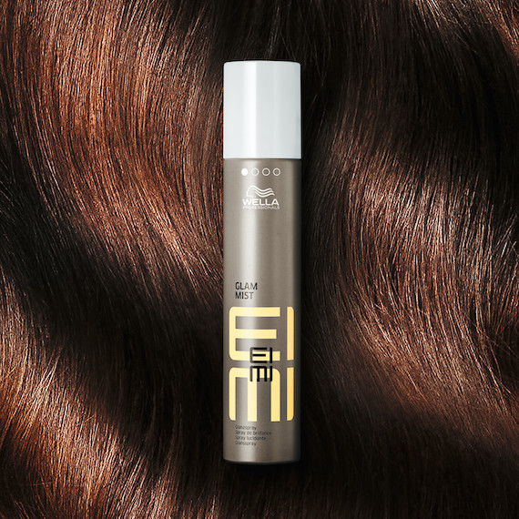 A bottle of EIMI Glam Mist in front of a model’s glossy, wavy, dark brown hair.