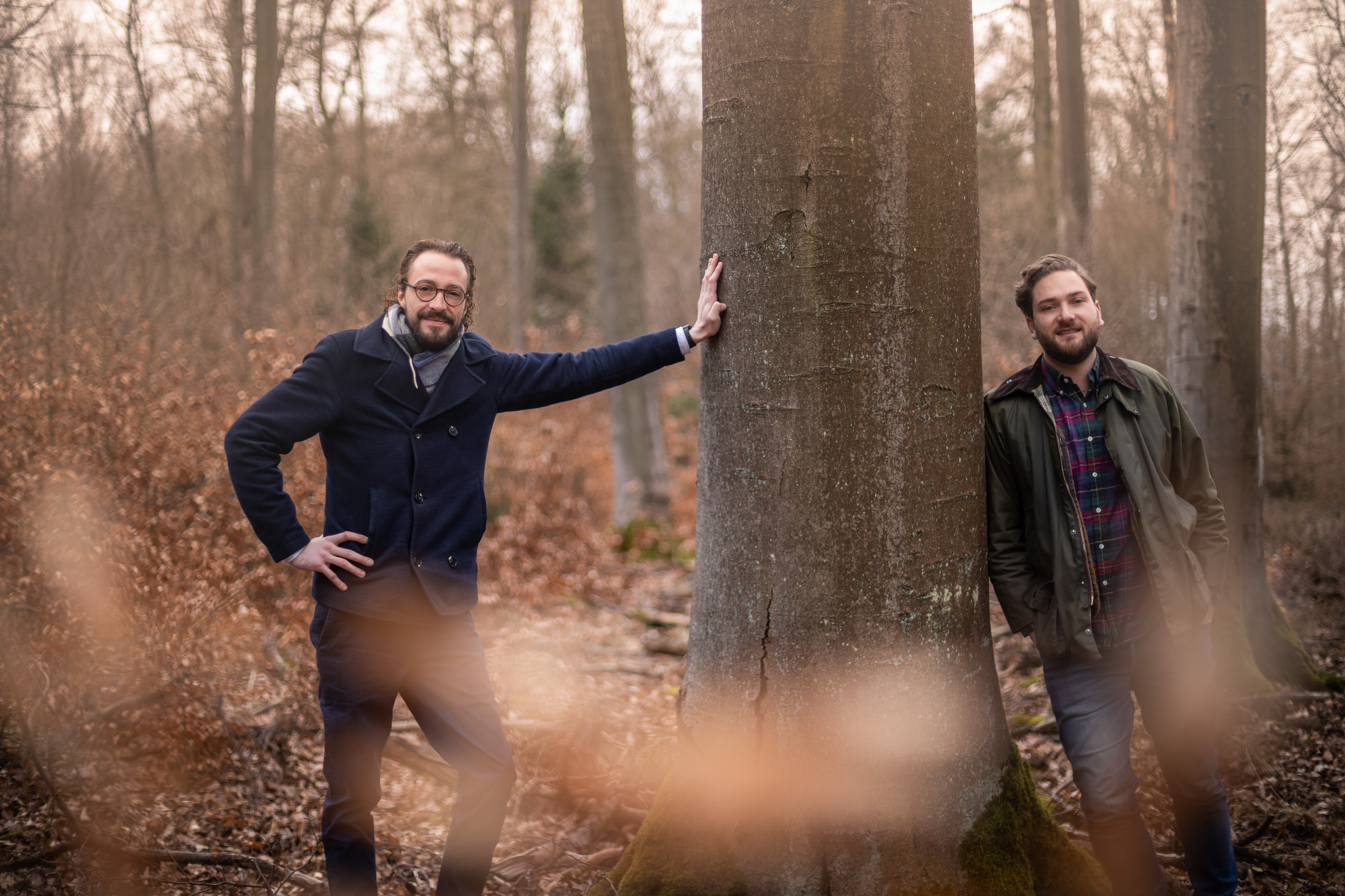 Zwei Männer stehen im Wald und lehnen an einem Baum.