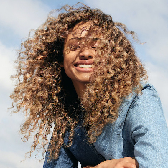 Woman with glossy, caramel blonde, curly hair smiles at the camera, created using Wella Shinefinity.