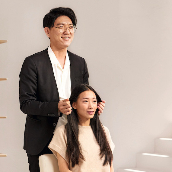 A hairstylist stands behind their client, who is sitting in a chair.