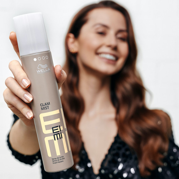 A model with long, wavy brown hair holds out a bottle of EIMI Glam Mist by Wella Professionals