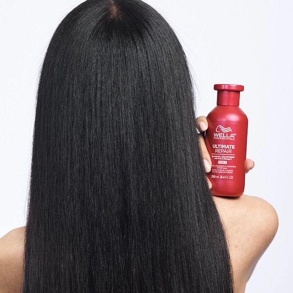 Ein Model mit langen, schwarzen Haaren steht mit dem Rücken zur Kamera und hält eine Flasche Wella Ultimate Repair Shampoo auf der Schulter