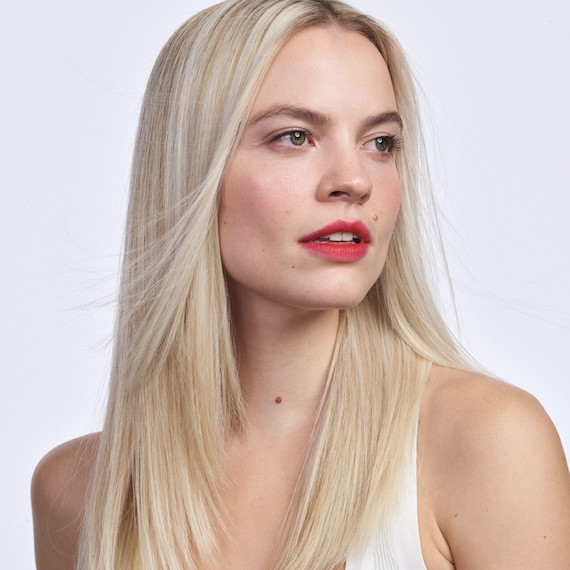 Headshot of a model with straight, platinum blonde hair wearing a white top and red lipstick