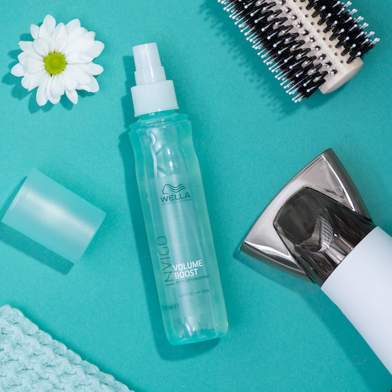 A blue bottle of Wella Professionals Invigo Volume Boost Hair Mist surrounded by a daisy flower, hairbrush and hairdryer 