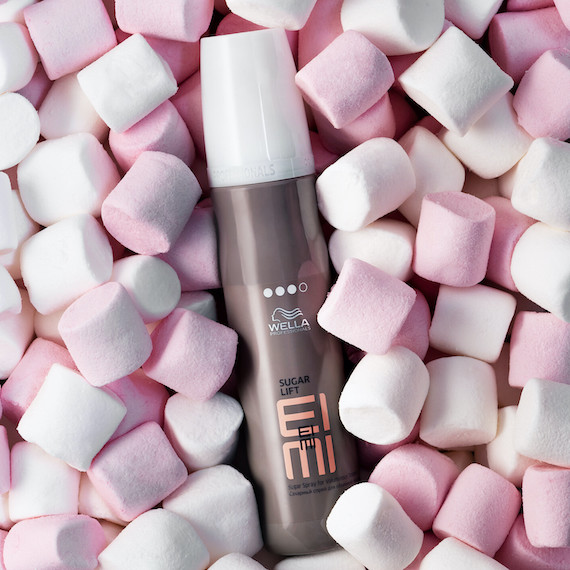 Eine graue Flasche Wella Professionals EIMI Sugar Lift Texturspray, umgeben von rosafarbenen und weißen Marshmallows