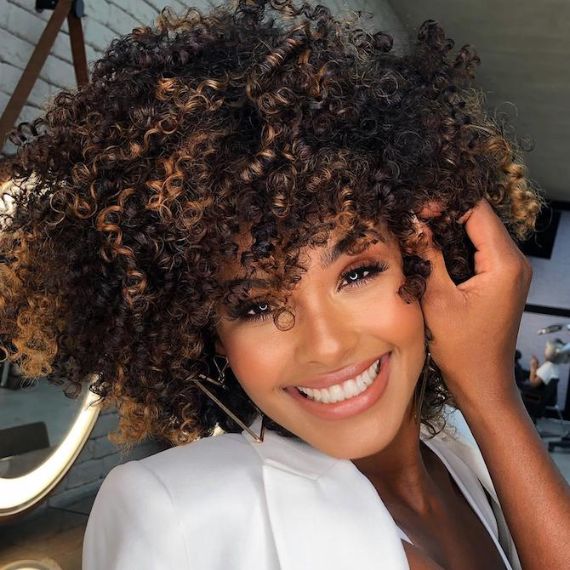 Woman smiles at the camera with curly, dark brown hair and chocolate balayage, created using Wella Professionals.