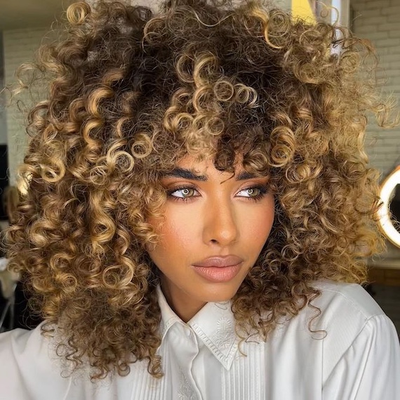 Model with curly, brown hair and glossy, golden blonde highlights.
