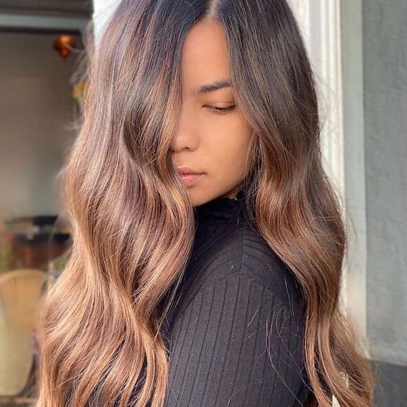 Model looks over their shoulder with their wavy, brown caramel ombre hair partially concealing their face