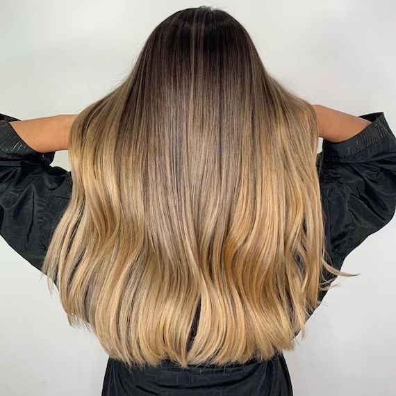 Model lifts their long, caramel brown to honey blonde ombre hair