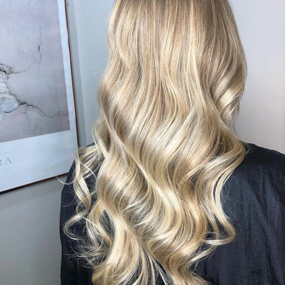 Image of the back of a woman’s head with long hair, wearing powdered blonde balayage color styled in tonged waves.