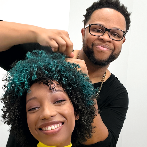 Model’s black, curly hair is being styled by a professional hairdresser. 