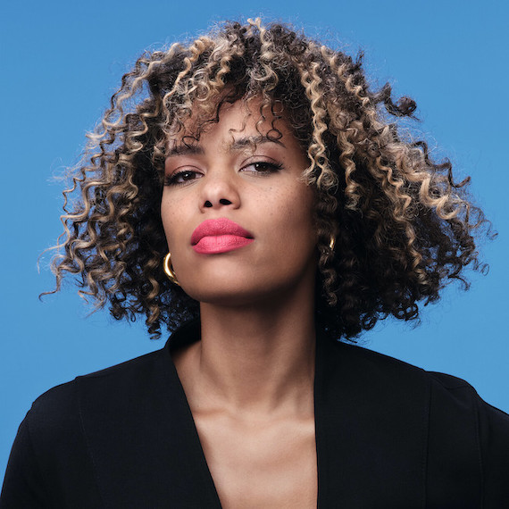 Model with dark brown, curly hair and chunky, honey blonde highlights looks directly into the camera. 