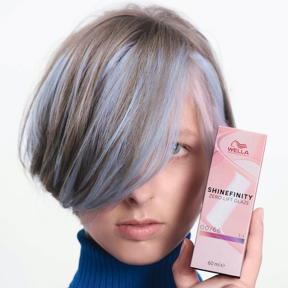 Model with short, platinum blonde hair and blue highlights holds up a box of Wella Shinefinity color.