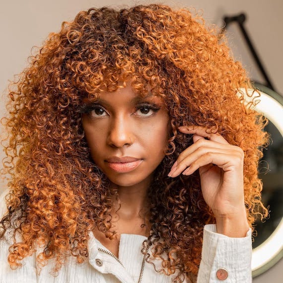 Model with long, shiny, curly hair featuring a brown root shadow and bright copper lengths.