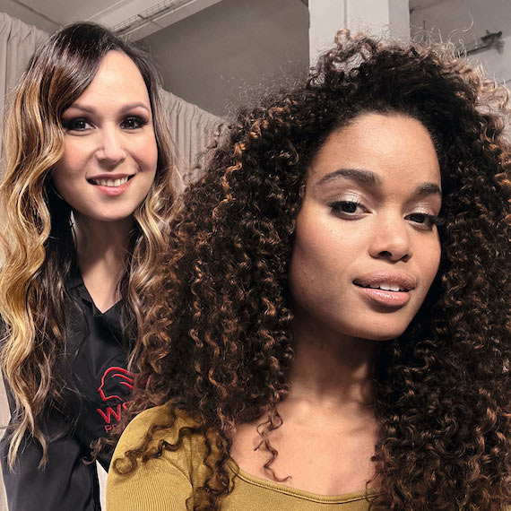 A hairdresser and a model with long, voluminous, curly hair smile directly into the camera.