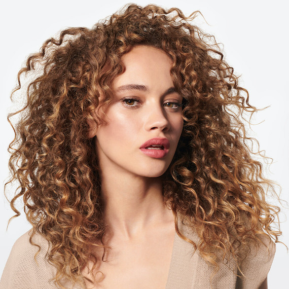 Model with long, curly, shiny, golden brown hair faces the camera.