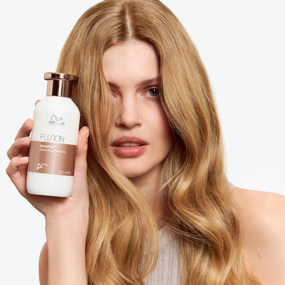 Model with long, honey blonde hair holds up a bottle of Fusion Intense Repair Shampoo.