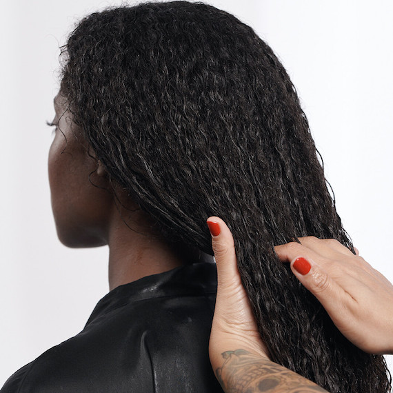 Hairdresser runs their hands through a model’s long, curly, black hair.