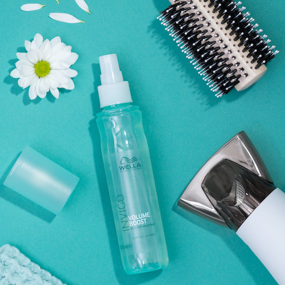 INVIGO Volume Boost Leave-In Spray on a green background surrounded by flowers, a round brush and a hairdryer.