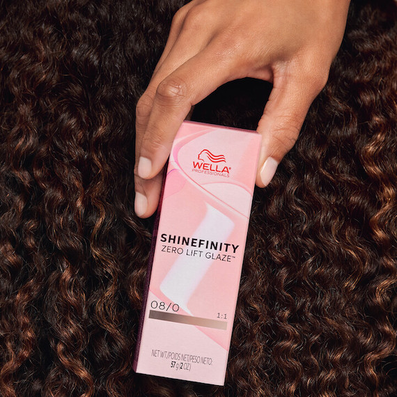 Model with curly, dark brown hair holds up a box of SHINEFINITY.