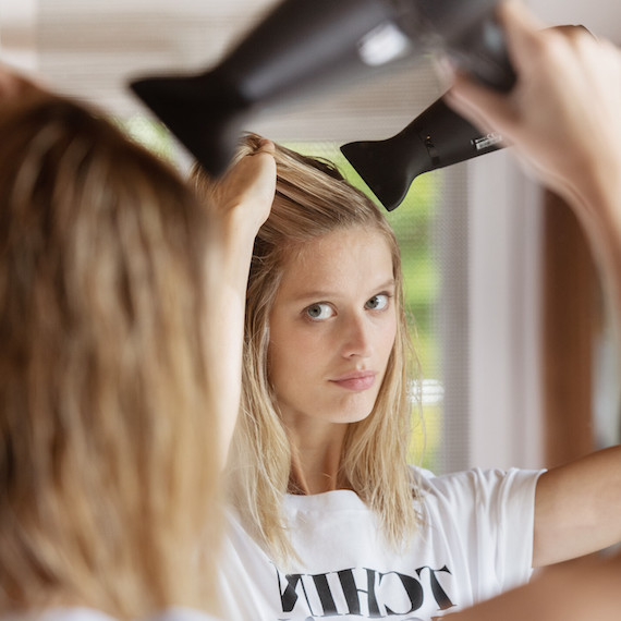 Blonde-haired model looks into mirror while blow-drying their hair.