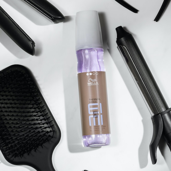 A bottle of EIMI Thermal Image is surrounded by a hairbrush, hair straighteners and a curling iron.