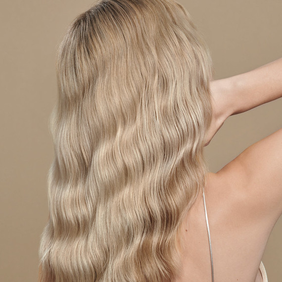 Model stood in front of a beige wall facing away from the camera. Their blonde hair is styled into beachy waves