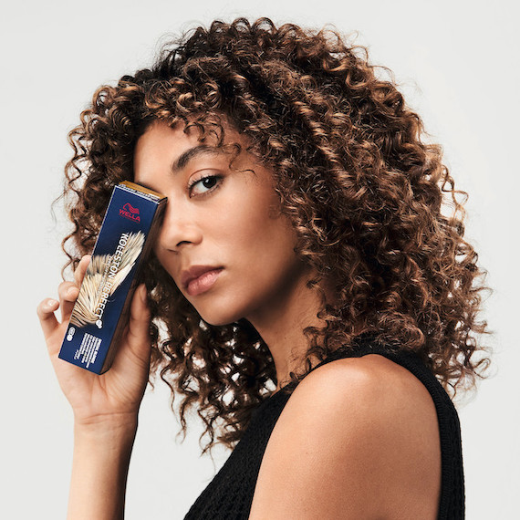 Model with highlighted curly hair looks over her shoulder holding a box of Koleston Perfect Special Blonde over one eye
