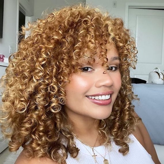 Model with honey blonde highlights through curly hair smiles at the camera.