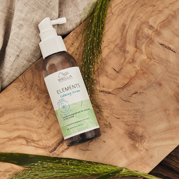A bottle of Elements Calming Serum sits on a wooden board surrounded by green vegetation and cream fabric