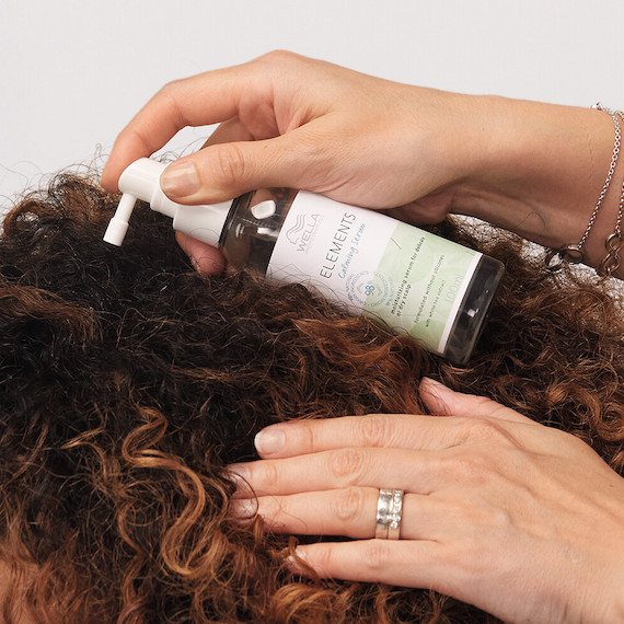 Close-up of hands resting on a head of curly hair while spritzing Elements Calming Serum by Wella Professionals on the roots