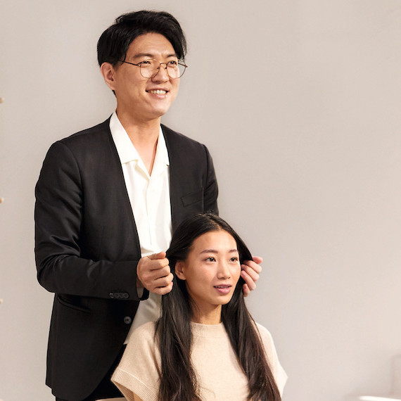 Hairstylist stands behind their client whilst holding and analyzing their dark brown hair