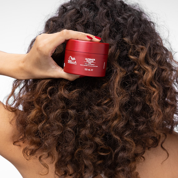Back of model’s head with long, dark brown, curly hair. They’re holding up the ULTIMATE REPAIR Mask.