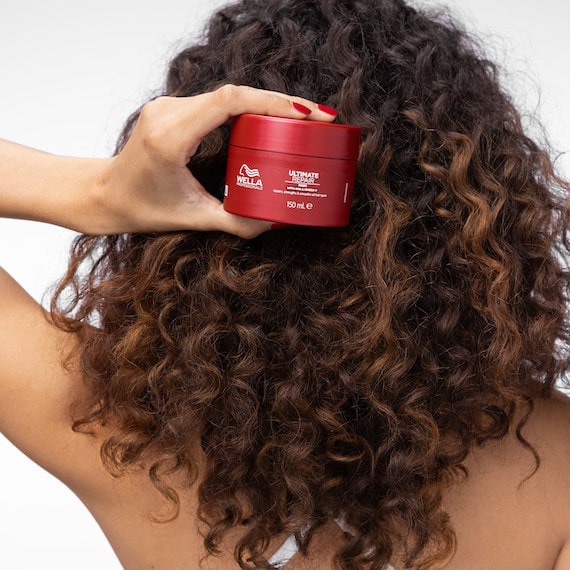 Back of model’s head with long, dark brown, curly hair, holding up the ULTIMATE REPAIR Mask.
