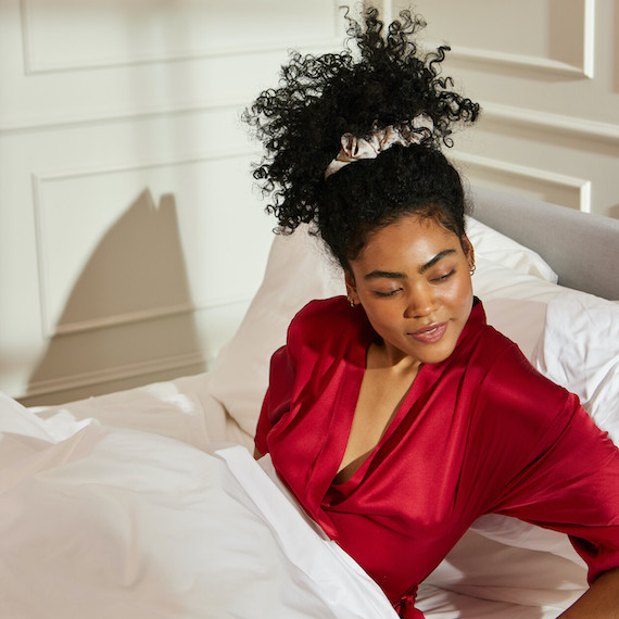 Model with dark brown, curly hair sits up in bed.