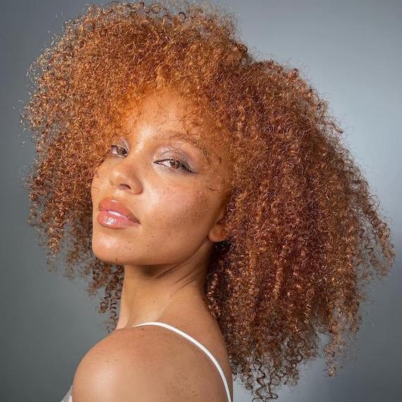 Headshot of a model with curly ginger brown hair stood in front of a gray backdrop 